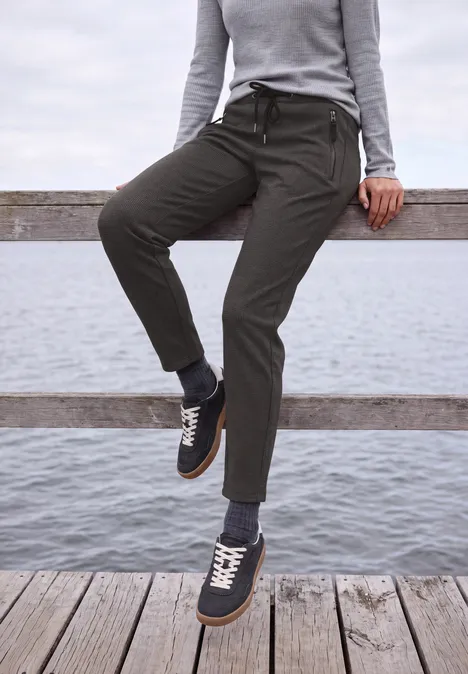 Mid Waist Barrel Leg Hose im Casual Fit creamy mocha Person lehnt an einem Holzgeländer am Wasser, trägt graue Stoffhose und dunkelblaue Sneaker.