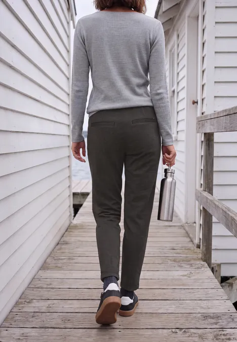 Mid Waist Barrel Leg Hose im Casual Fit creamy mocha Person von hinten in grauem Pullover und Hose, die mit einer Flasche in der Hand über einen Holzsteg an Häusern geht.