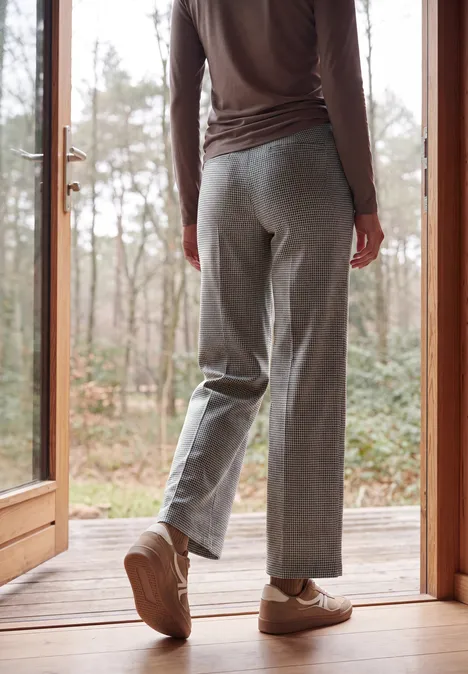 Person steht auf einer Holzterrasse vor einer Glastür, trägt graue Hose und beigen Sneaker, im Hintergrund Bäume.