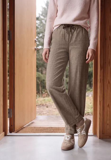 Person steht in einem Holztürrahmen, trägt rosa Pullover, karierte Hosen und beiger Sneaker, Natur im Hintergrund.