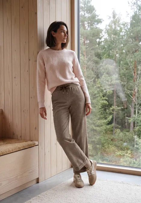 Eine Frau in hellem Pullover lehnt entspannt am Fensterrahmen, mit Blick auf den bewaldeten Garten.