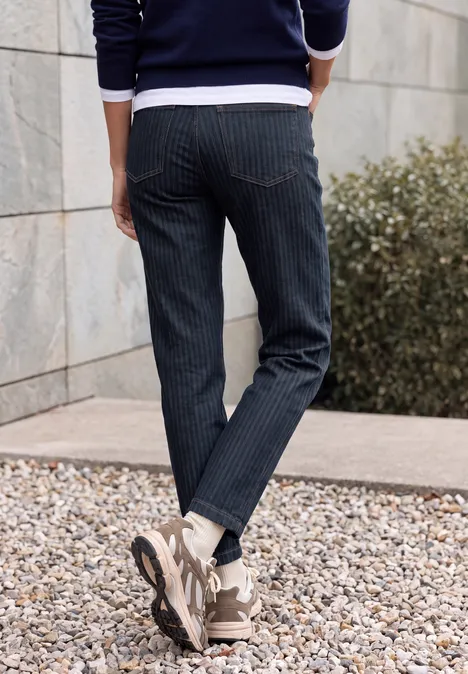High Waist Barrel Leg Jeansim Loose Fit rinsed wash Person steht im Freien, trägt dunkelblaue Nadelstreifenhose und Sneaker auf Kiesboden vor einer Mauer.