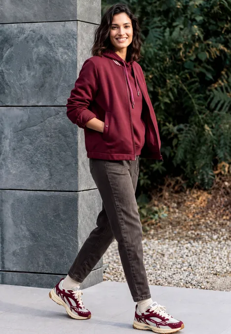 Jeans met wijd uitlopende pijpen in garment-dye-look truffle brown Een lachende vrouw in een bordeauxrode hoodie, bruine broek en gekleurde sneakers leunt ontspannen tegen een muur buiten.