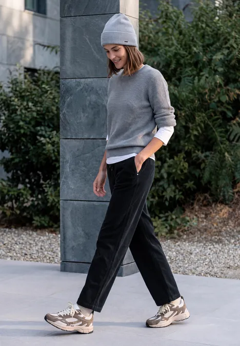 Frau in grauem Pullover, schwarzer Hose und Mütze spaziert lächelnd an einer Fassade entlang.