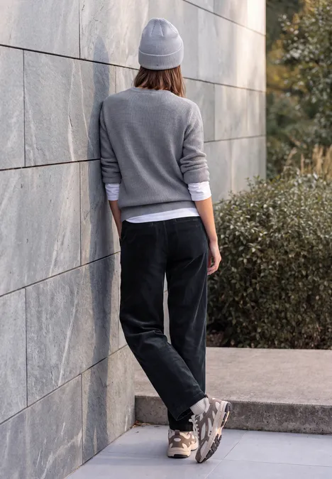 Person mit grauer Mütze und Sweatshirt lehnt an Steinmauer, trägt schwarze Hose und Sneaker.