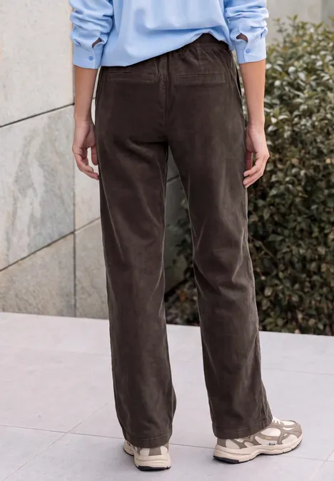 Personne vue de dos porte un pantalon en velours brun et des baskets beiges devant un mur