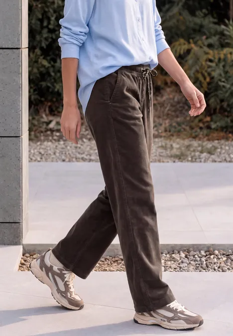 Personne en pantalon en velours côtelé marron, chemise bleu clair et baskets beige sur un trottoir.