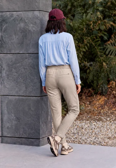 Person mit roter Kappe, hellblauer Bluse und beigen Hosen läuft an Mauer und herbstlichem Laub vorbei.