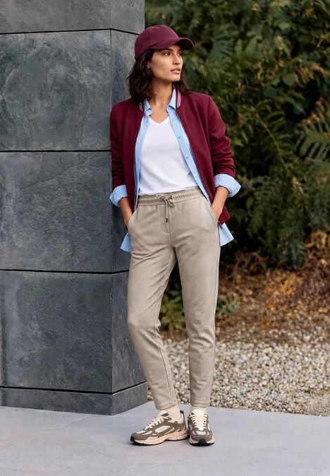 Frau lehnt an Hauswand im Freien, trägt burgunderrote Jacke, hellblaue Bluse, weißes Top, beige Hose und Sneaker.