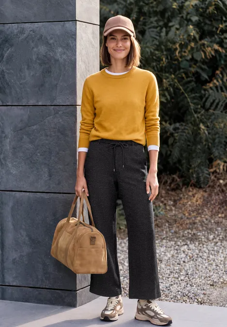 Frau mit gelbem Pullover, schwarzer weiter Hose, Schlangenprint-Stiefeln und Tasche steht vor moderner Fassade im Freien.