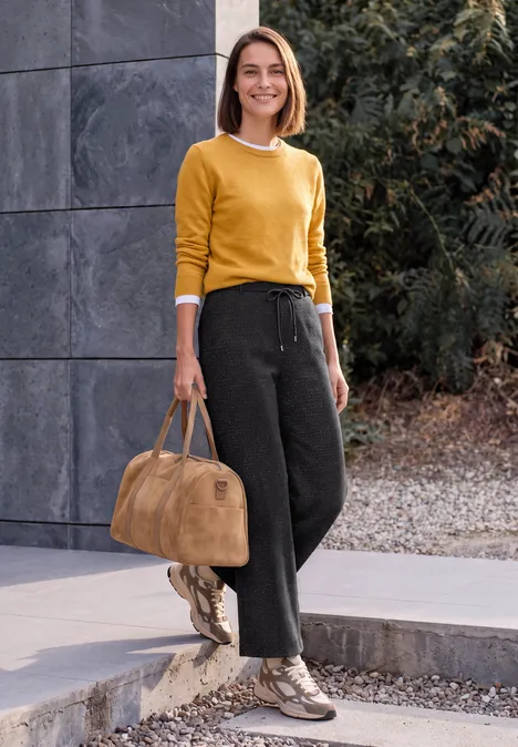 Frau in gelbem Pullover und schwarzer Hose mit beiger Tasche, draußen stehend vor einer grauen Betonwand und Grün.