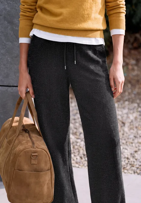 Eine Person trägt schwarze Hose und gelben Pullover und hält eine braune Wildledertasche.