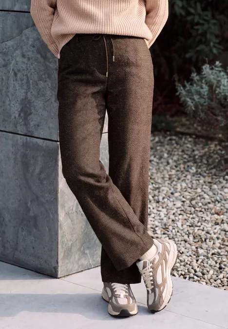 High Waist Wide Leg Hose im Loose Fit truffle brown Eine Person trägt braune, weite Hosen und weiße Sneaker, steht auf Kies neben einer Steinwand.
