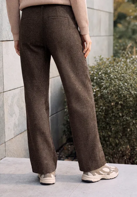 High Waist Wide Leg Hose im Loose Fit truffle brown Person trägt braune weite Hosen und weiße Sneaker, steht vor einer Steinmauer neben Büschen.