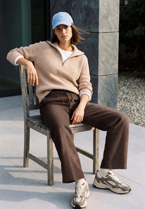 High Waist Wide Leg Hose im Loose Fit truffle brown Frau sitzt lässig auf Stuhl im Freien, trägt Pullover, weite Hose, Sneaker und Cap.