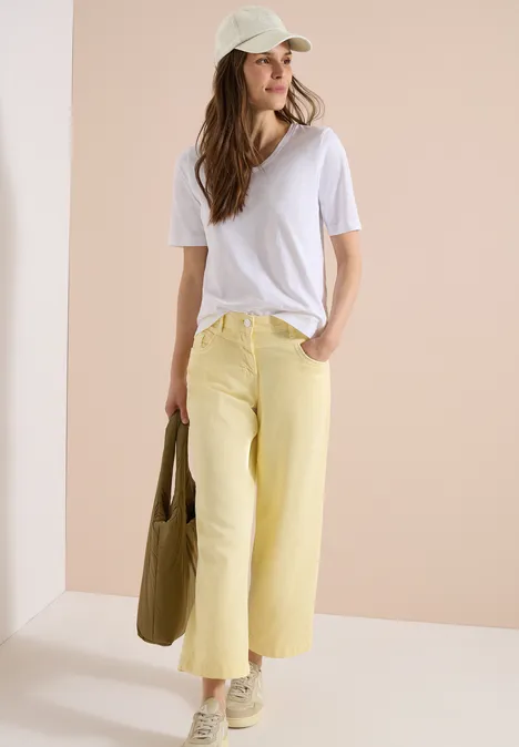 Frau in weißem T-Shirt, pastellgelben Hosen, Baseballcap und grünem Beutel vor hellem Hintergrund.
