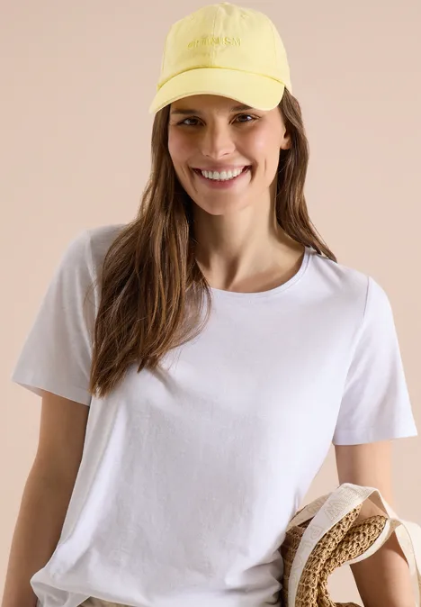 Frau in weißem T-Shirt und gelber Baseballkappe, hält einen Strohhut vor neutralem Hintergrund.