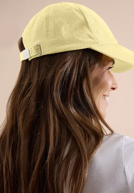 Rückansicht einer Person mit langen braunen Haaren und gelbem Baseballcap vor beigem Hintergrund.