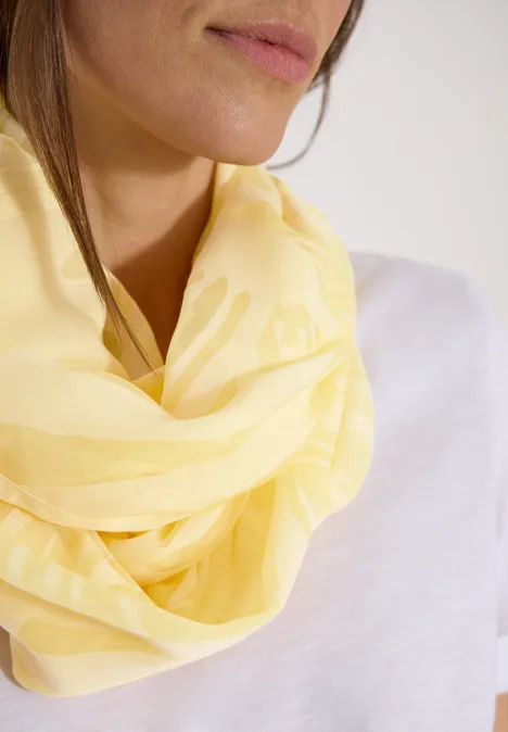 Eine Person trägt einen hellgelben Schal und ein weißes Shirt, nur Hals und Unterkiefer sind sichtbar.