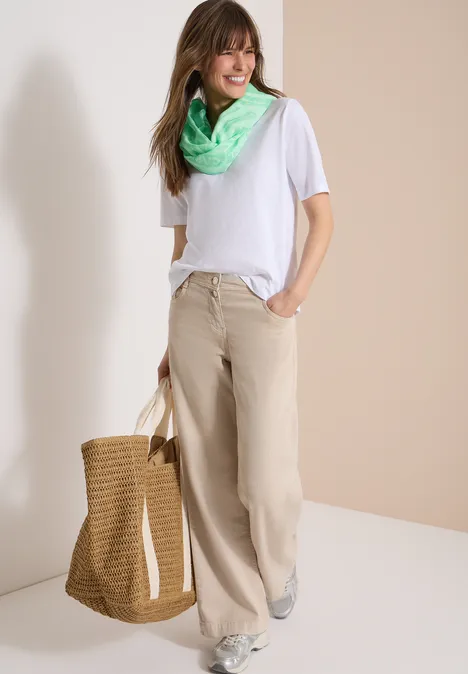 Frau in weißem T-Shirt, beiger, weiter Hose, mit grünem Halstuch, trägt eine Stroh-Tasche.