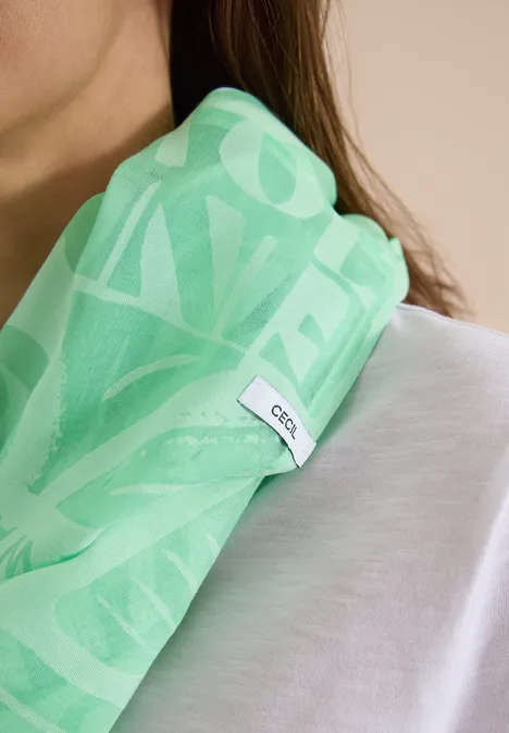 Hellgrüner Schal mit weißem Muster und GCDS-Label, lässig über eine Schulter gelegt, kombiniert mit weißem Shirt.