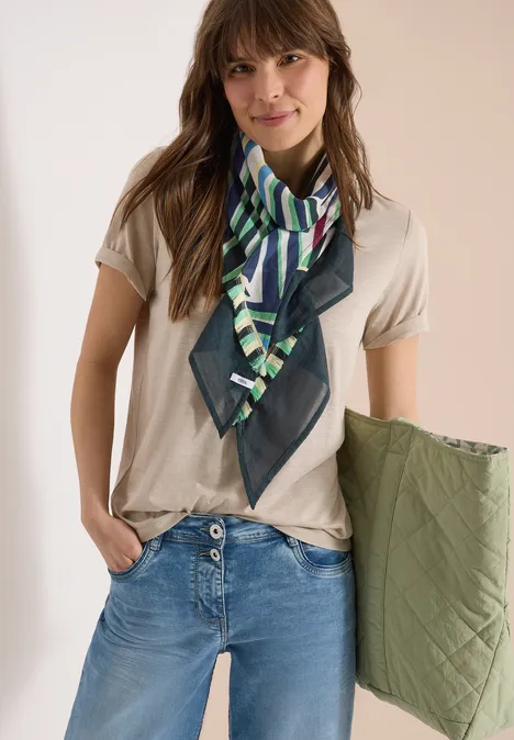 Frau in beigem T-Shirt, blauer Jeans, grün gemustertem Seidenschal, hält olivgrüne gesteppte Jacke