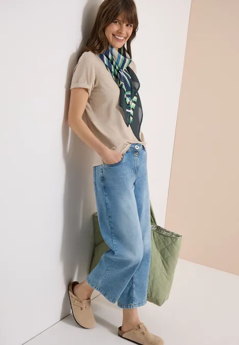 Eine Frau lehnt an einer Wand, trägt weite Jeans, ein beiges Top, grünen Schal und grüne Tasche.