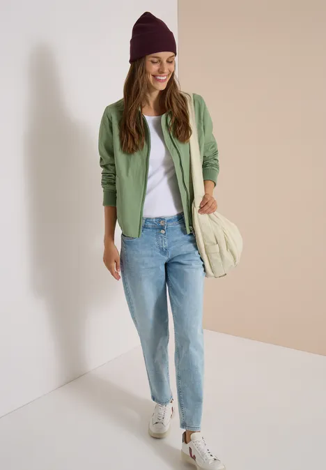 Jeune femme en gilet vert, T-shirt blanc, jean, bonnet violet et baskets sourit dans une pièce claire.