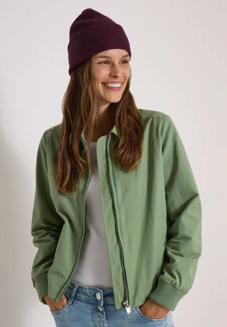 Femme souriante en veste verte, t-shirt gris et bonnet violet sur fond neutre.
