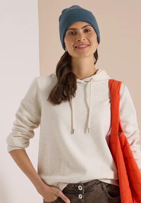 Femme souriante en sweat clair, bonnet bleu et veste rouge posée sur l’épaule.