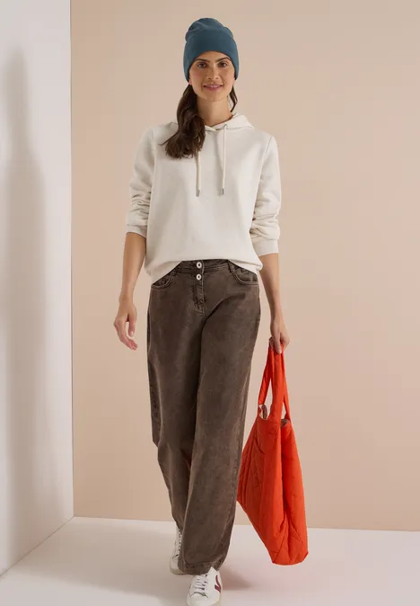 Femme en sweat à capuche blanc, jean marron ample, bonnet bleu et sac rouge devant un fond neutre.