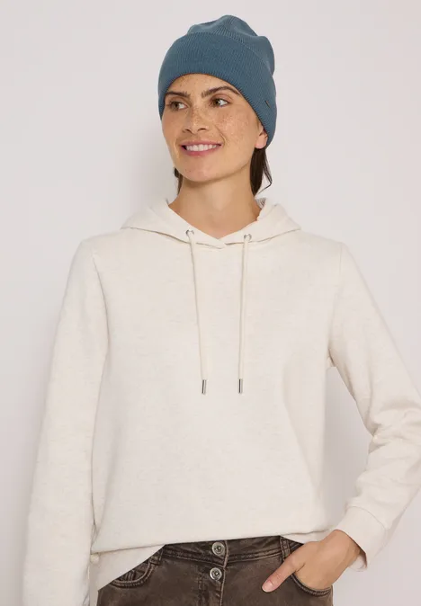 Femme portant un sweat à capuche beige et un bonnet gris-bleu, souriant devant un fond neutre.