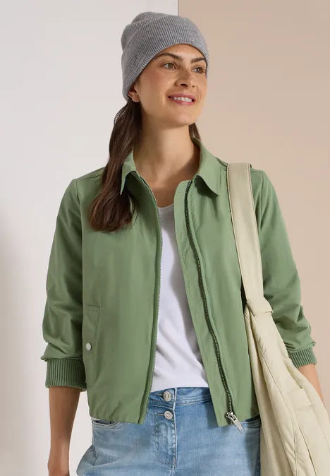 Jeune femme devant un fond clair, portant un bonnet gris, une veste verte, un t-shirt blanc et un sac en toile.