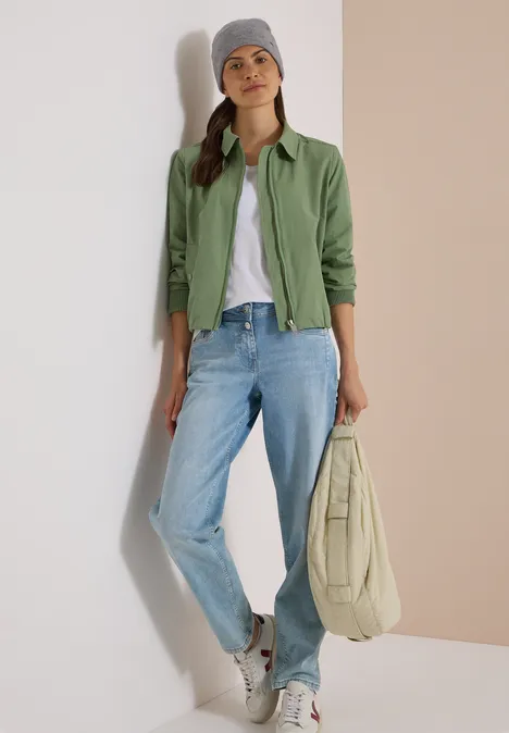 Femme en veste verte, t-shirt blanc, jean bleu et bonnet gris, tenant un sac beige.