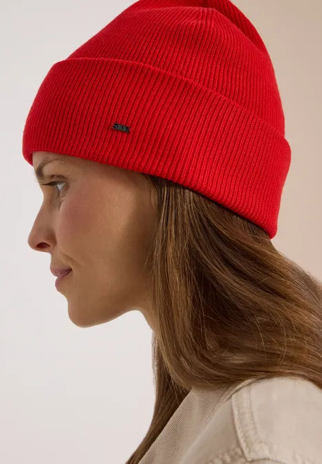 Profil latéral d’une femme portant un bonnet rouge et une veste claire sur fond neutre.
