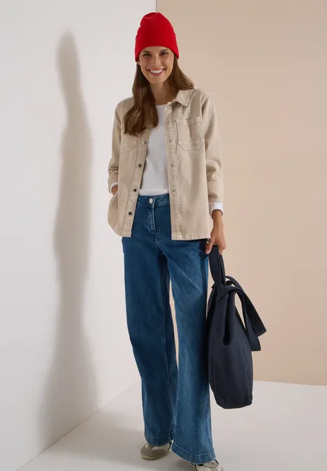 Femme souriante en bonnet rouge, veste beige, pull blanc et jean large, avec un sac bleu.