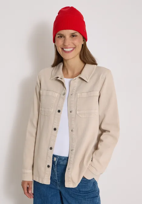 Jeune femme souriante en bonnet rouge, veste beige et t-shirt blanc sur fond clair.