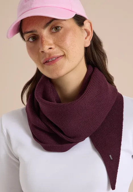 Femme portant une casquette rose, une écharpe violette et un haut blanc sur fond neutre.