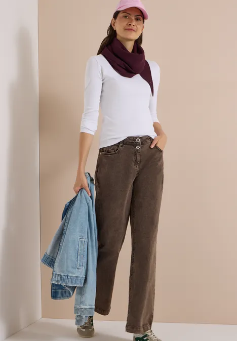 Femme en chemise blanche, pantalon marron, casquette rose, tenant une veste en jean, fond neutre.
