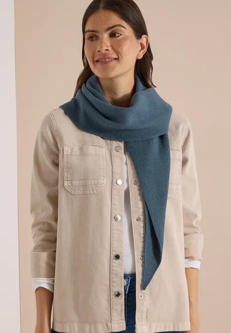 Femme en veste claire et écharpe bleue, souriante devant un fond neutre.