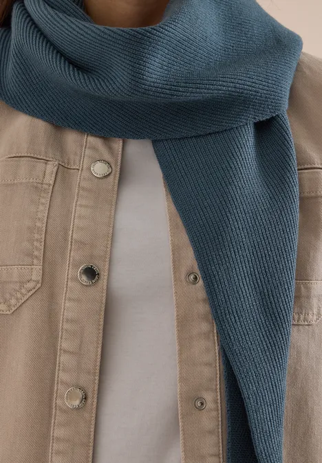 Écharpe en tricot bleu autour du cou, veste beige clair et t-shirt blanc.