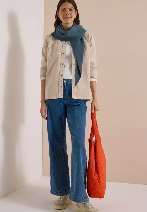 Femme avec veste claire, top blanc, jean large bleu, écharpe bleue et sac orange sur fond neutre.