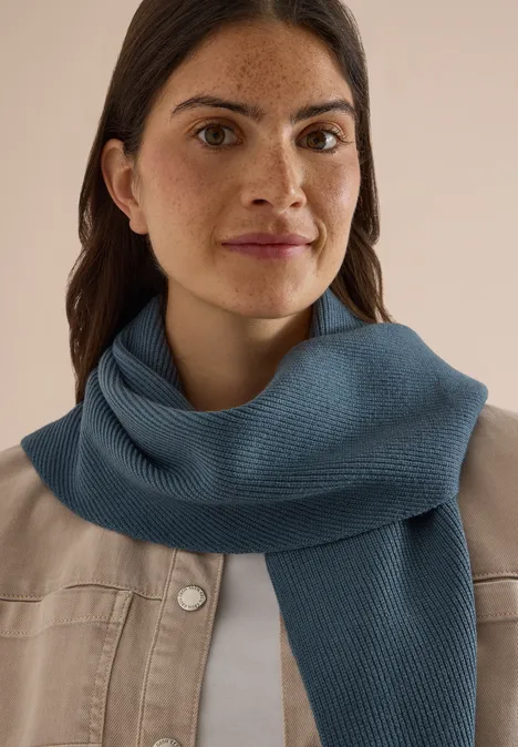 Femme aux cheveux bruns en veste claire portant une écharpe bleue, regardant l'appareil photo avec un léger sourire.