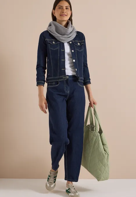 Femme en veste et pantalon en jean bleu foncé, écharpe grise, baskets blanches et grand sac vert.