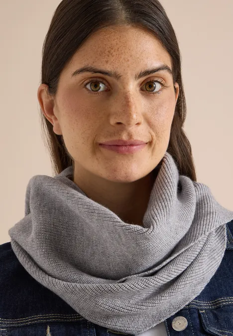 Femme aux cheveux bruns portant une écharpe grise et une veste en denim sur fond clair
