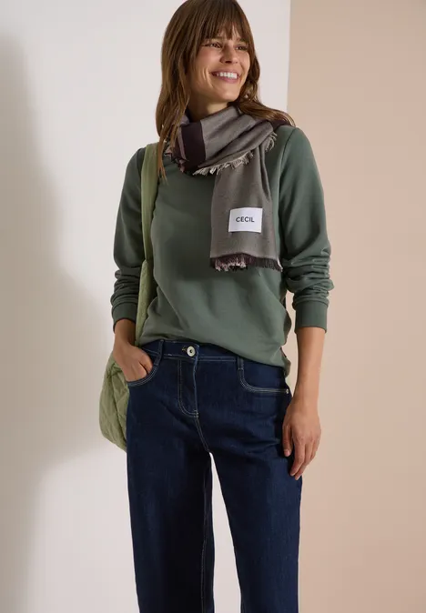 Femme portant un pull vert, une écharpe, un jean et un sac, souriante devant un fond neutre.