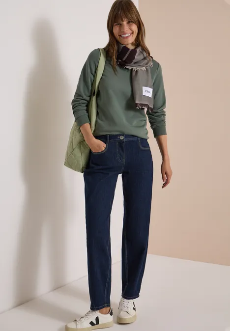 Une femme souriante porte un pull vert, un jean, une écharpe, un sac et des baskets blanches sur un fond neutre.