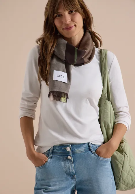 Jeune femme souriante en t-shirt blanc, jean, écharpe à carreaux et sac vert.