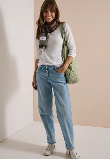 Jeune femme souriante en chemisier blanc, jean ample, écharpe et sac vert devant un mur neutre.