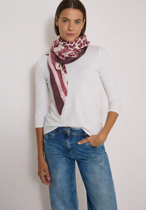 Foulard triangulaire à motif léopard berry red Frau in weißem Langarmshirt, blauen Jeans und gemustertem Schal steht mit einer Hand in der Hosentasche.